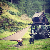 Off-road vehicle with rooftop tent, outdoor tarp, and a dog near a campfire in a forested area.